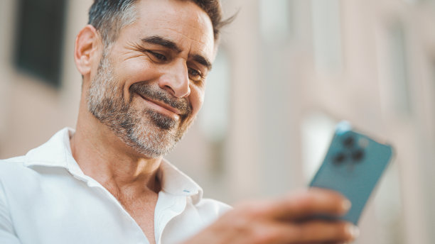 留着整齐胡须的成熟商人坐在城市金融区的长椅上使用手机。成功的男人在智能手机上滚动信息并微笑图片下载