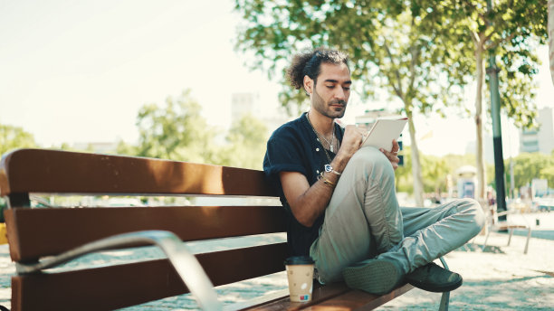 年轻的意大利小伙子扎着马尾和胡茬坐在街上的长凳上，在纸上用笔画草图图片下载