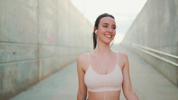 年轻的运动女子长马尾穿着米色运动上衣有线耳机，微笑着走着图片下载