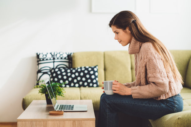 侧视图一个女人坐在沙发上的笔记本电脑上工作，而她的早上咖啡图片下载