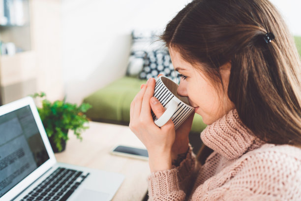 近距离女子喝茶从一个杯子，而在家里工作在她的笔记本电脑图片下载