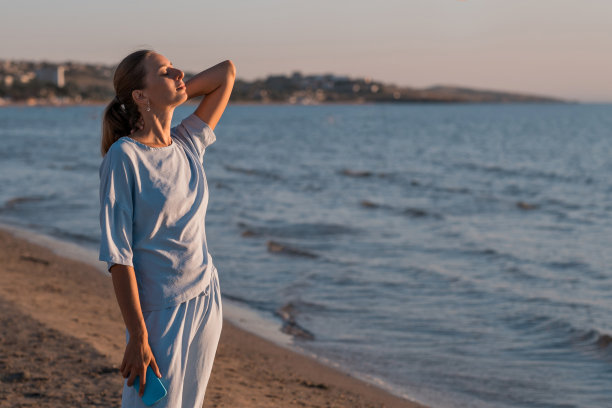 一个金发碧眼的女孩在夕阳下以大海为背景。在海滩上在线工作。图片下载