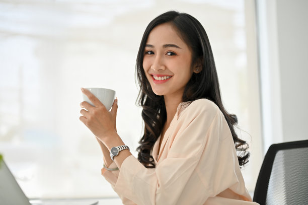 迷人的亚洲女商人坐在她的办公桌前，拿着咖啡杯，微笑着看着相机图片下载