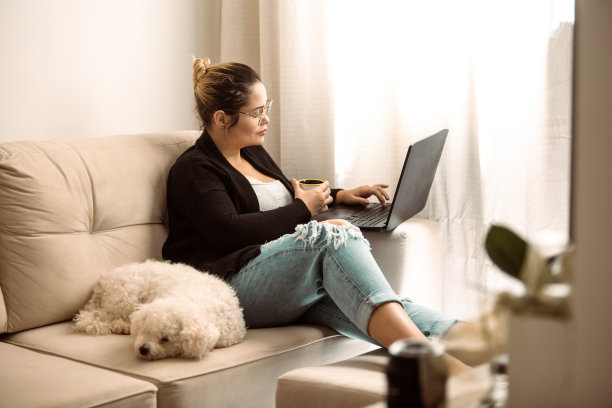 年轻女子喝咖啡，同时使用她的笔记本电脑在家里工作图片下载