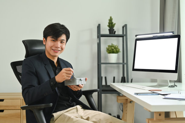 微笑的亚洲男性员工坐在办公桌前，电脑屏幕空白，在办公室享受午休时间。图片下载