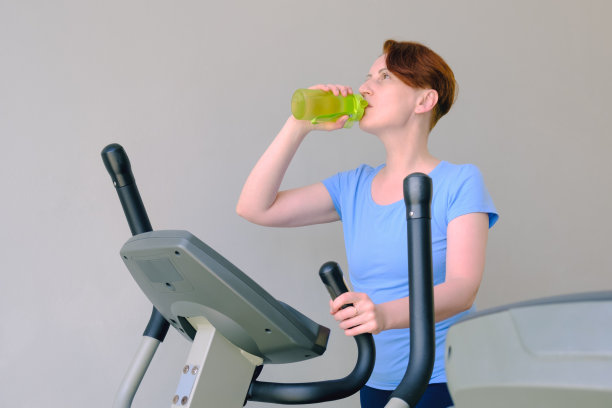 一名女子站在健身房的跑步机上，从运动瓶子里喝水图片下载