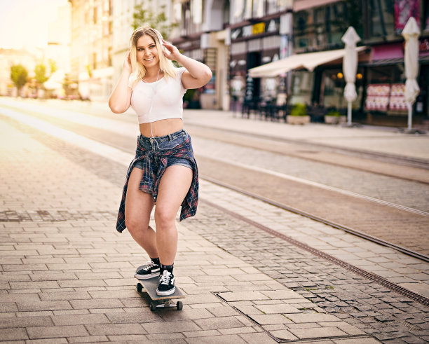 快乐的少年女孩在城市街道上滑板，同时听音乐耳机-保持微笑和享受生活的概念-背光夏日生活方式的照片图片下载