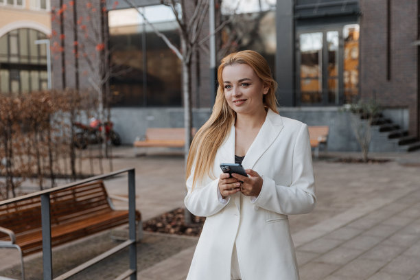 金发时尚的女人在超大的白色夹克上使用手机在现代城市的背景图片下载