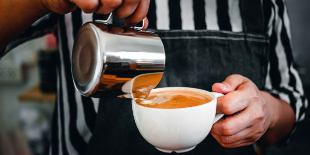 特写的手，一个专业的咖啡师在咖啡店把蒸牛奶倒进咖啡杯制作拿铁菜单图片下载