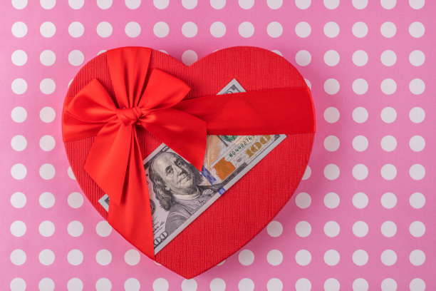 礼盒在一个人的心脏符号的形式与100美元的粉红色背景钞票图片下载