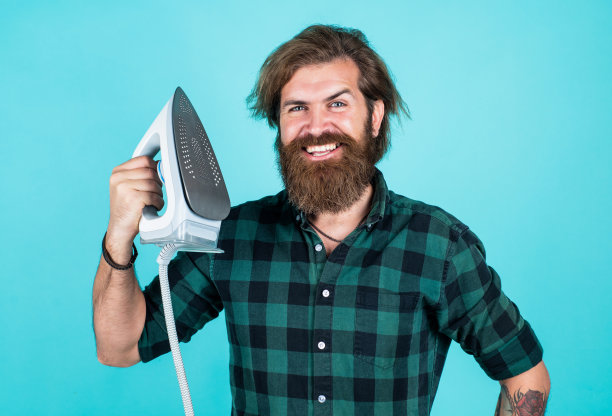 好的质量。家庭家务。成熟粗暴的潮人使用现代科技。家庭的概念。小伙子勤勤恳恳地熨衣服。家用电器。快乐的大胡子男人拿着热气腾腾的熨斗图片下载