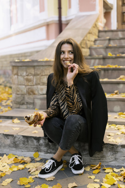 美丽的黑发女孩坐在楼梯上，掰下一块，吃了一个羊角面包。一个女孩正坐在一座老建筑附近的街上。图片下载