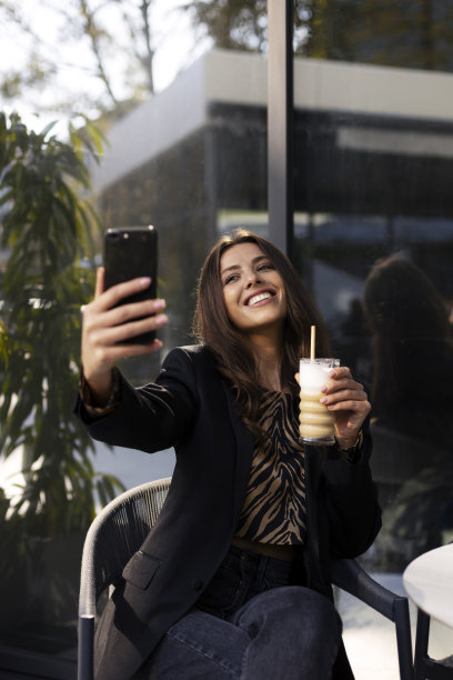 这名美丽的年轻女子一手拿着咖啡，一手拿着智能手机，端着一杯饮料，在户外的咖啡馆里自拍，并为她的粉丝拍摄了一段视频。图片下载
