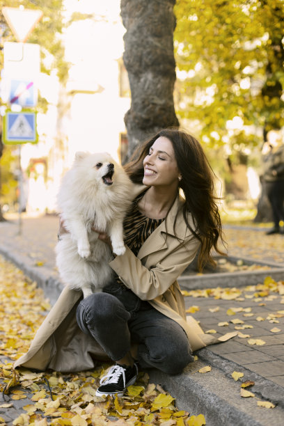 迷人的女孩抱着一只白色的狗，狗在叫，女孩看着他。带着宠物在户外散步。图片下载