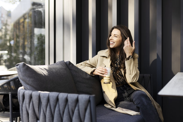 积极，微笑的年轻女子坐在沙发上，在咖啡馆的露台上喝着饮料。迷人的女孩休息，看向别处。图片下载