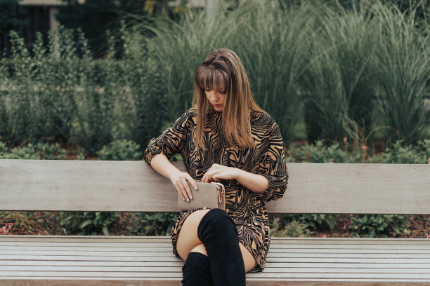 年轻女子坐在长凳上，穿着裙子，看着她的包/钱包图片下载