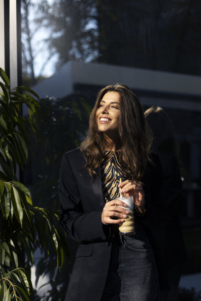 迷人的年轻女子站在咖啡在她的手中对着玻璃的背景，现代建筑。一个微笑的黑发女孩抬头，阳光正照耀着她。图片下载