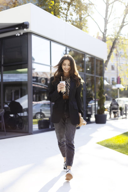 快乐的年轻女孩手里拿着饮料，走在大街上，背景是一座现代建筑。迷人的女人在休闲风格。图片下载