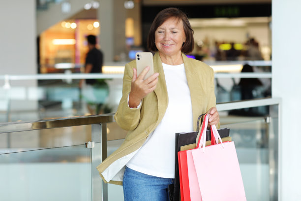 在购物中心内的中年妇女正在使用智能手机并拿着装满商品的彩色包裹图片下载
