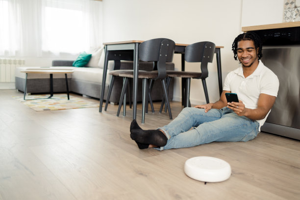 年轻的非洲裔美国人正在用智能手机调整他的智能吸尘器图片下载