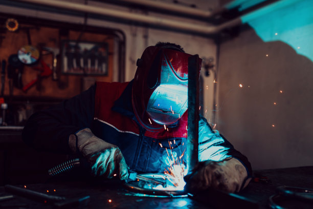 在工业车间，专业焊工穿着防护服，戴着面罩，与其他工人一起在工业台上焊接金属管道。图片下载