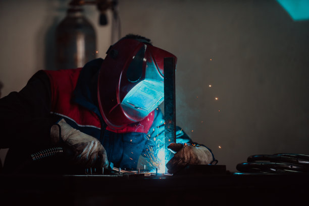 在工业车间，专业焊工穿着防护服，戴着面罩，与其他工人一起在工业台上焊接金属管道。图片下载