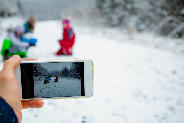 男性手持智能手机，拍孩子们在雪地里玩耍的照片。图片下载