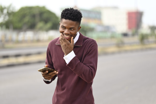有吸引力的年轻非洲裔美国人在街上拿着他的智能手机，微笑着快乐的笑表情，用他的手来演示他的下巴图片下载