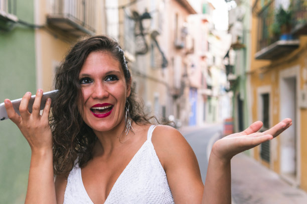 黑发女子拿着她的手机靠近她的耳朵听音频信息，在她的脸上惊讶的表情，她走在五颜六色的街道，阿利坎特，西班牙。图片下载