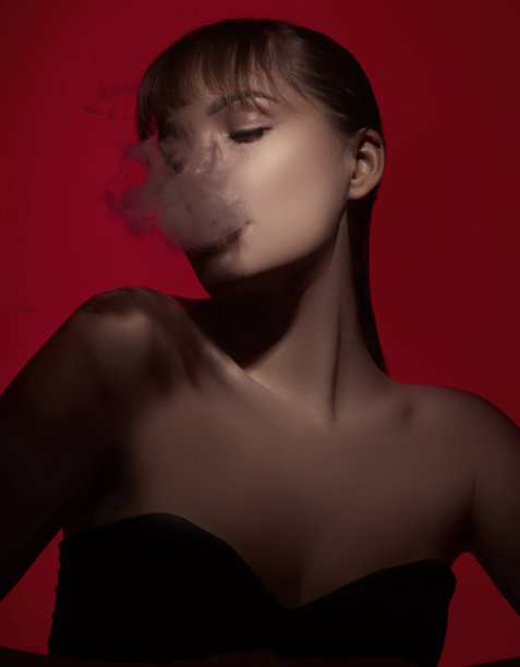 美丽的女人的肖像闭上眼睛，同时从她的嘴吹烟在戏剧性的灯光下在红色背景前。图片下载
