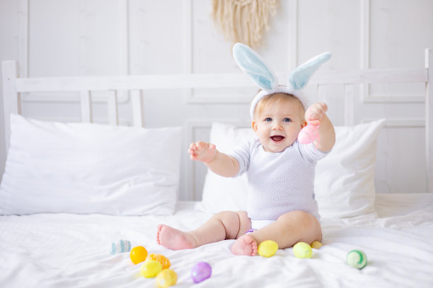 可爱的小男孩带着兔子耳朵和彩色的鸡蛋在家里的白色床上玩或吃鸡蛋，小金发宝宝，快乐的复活节概念图片下载