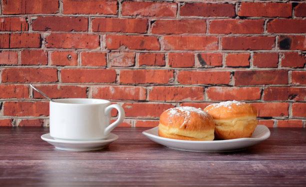 红砖墙边的木桌上放着一杯热黑咖啡和一个盘子，盘子里有两个新鲜的甜甜圈，里面有糖粉。图片下载