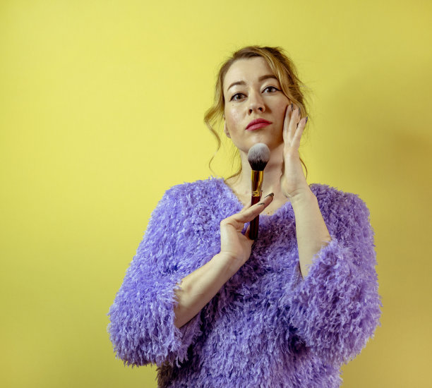 一个穿着蓬松紫色毛衣的年轻女子拿着化妆刷靠近下巴，背景是孤立的黄色。美女在化妆。女人图片下载
