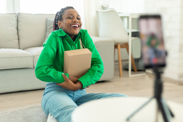 快乐的黑人妇女抱着纸箱，看着智能手机摄像头，在客厅为vlog录制视频图片下载