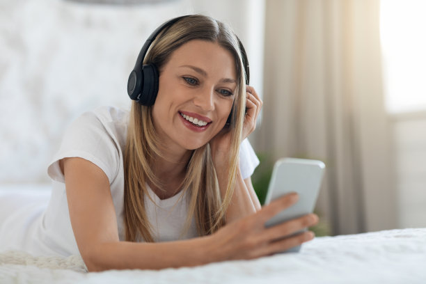 快乐的年轻女子躺在床上与智能手机和耳机图片下载