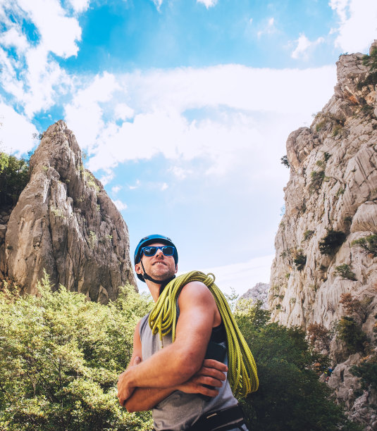 微笑的登山者肖像，戴着防护头盔和太阳镜，肩膀上挂着攀岩绳，站在Paklenica公园的岩石悬崖之间。积极的极限运动时间消费概念。图片下载
