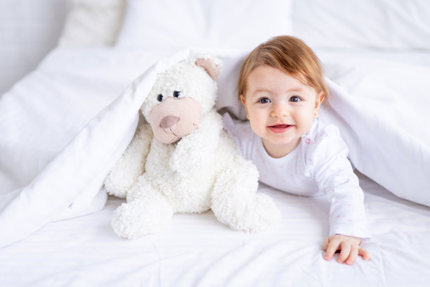 有趣快乐的小女孩穿着白色的衣服从床上的毯子下往外看，手里拿着泰迪熊，微笑着，一个小孩子在家里的棉床上睡了一觉后醒来图片下载