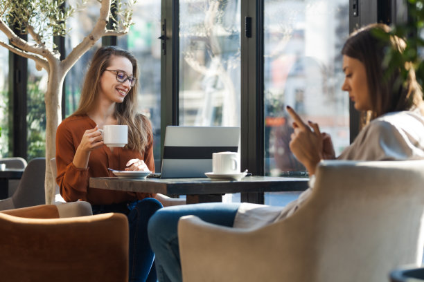 两个年轻的女商人在咖啡馆里一对一地开会。下班后的朋友们在阳光明媚的日子里闲聊，坐在靠窗的桌子上喝咖啡。图片下载