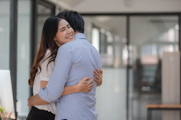 迷人的微笑的亚洲女人拥抱男人在室内。美丽的成功夫妇拥抱职场。丈夫和妻子拥抱在一起的概念。图片下载