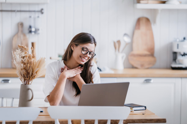 可爱的女学生在白色t恤坐在厨房桌子上的笔记本电脑，双手放在胸前，在修女课后表示感谢。远程教育。漂亮的意大利女人在家打视频电话。图片下载