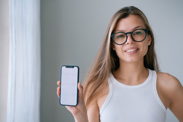 漂亮的白人女孩在眼镜显示手机与白色屏幕咧嘴笑在家里。欢快的西班牙年轻女子满意的新应用程序，演示智能手机。设备、技术。图片下载