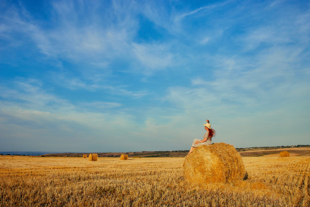 一位戴着帽子、穿着夏装的美丽年轻女子正坐在田野里的一捆干草上。乡村自然，麦田，休养在乡村，与自然合一图片下载