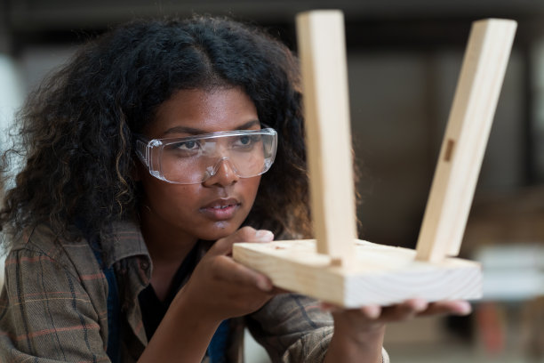 年轻的女木匠在木制车间制作椅子。黑女细木工戴着安全眼镜，在木制仓库的车间里坐着椅子。中小企业或小型企业的概念图片下载