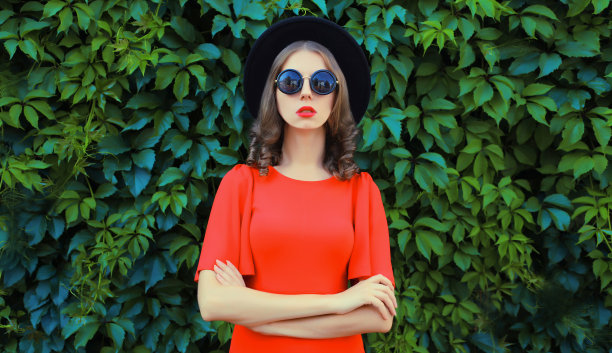 美丽的年轻女子的肖像摆着红色的连衣裙，黑色的圆帽子在绿叶墙的背景图片下载