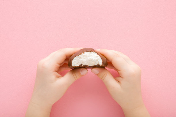 小女孩的手咬着白色香草软西风覆盖着深棕色巧克力釉在浅粉红色的桌子背景。柔和的颜色。特写镜头。孩子们吃甜食。自顶向下视图。图片下载