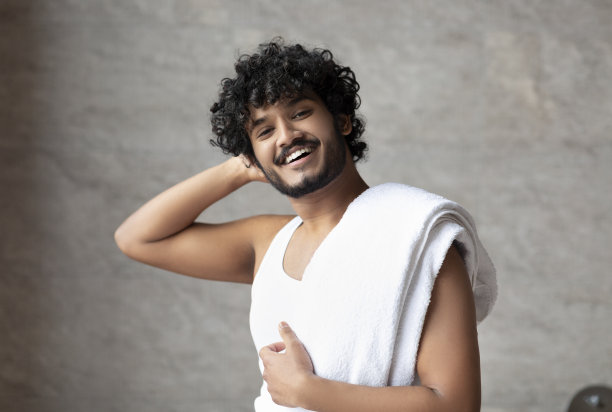 男性美容护理。快乐的印度男人肩上扛着毛巾站在浴室里，对着镜头微笑图片下载