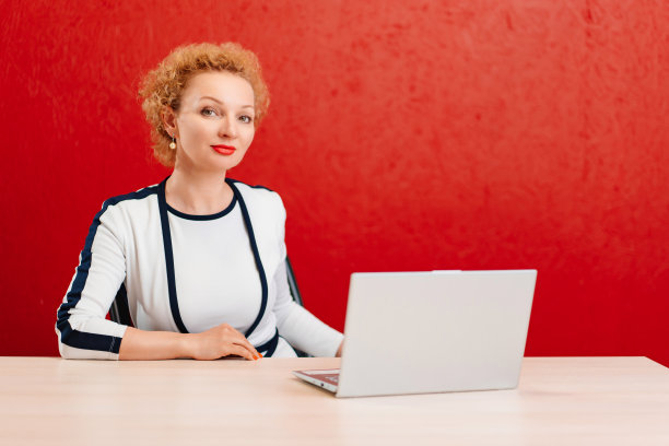 穿着黑白西装的金发女郎坐在书桌前，笔记本电脑靠在红墙上图片下载