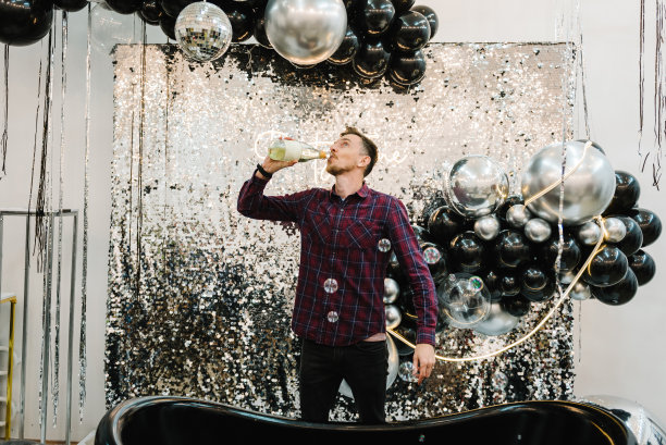 快乐男子喝香槟庆祝2023年。祝贺生日的地方。拱门或照相亭装饰黑色银色气球。墙，带浴室的摄影区。派对时间。图片下载