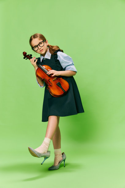 可爱的小女孩，才华横溢的音乐家穿着巨大的母亲的高跟鞋拉小提琴，有乐趣孤立在绿色的背景。教育、音乐、经典艺术、新视野、趣味理念图片下载