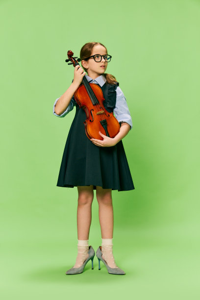 可爱的小女孩，才华横溢的音乐家穿着巨大的母亲的高跟鞋拉小提琴，有乐趣孤立在绿色的背景。教育、音乐、经典艺术、新视野、趣味理念图片下载